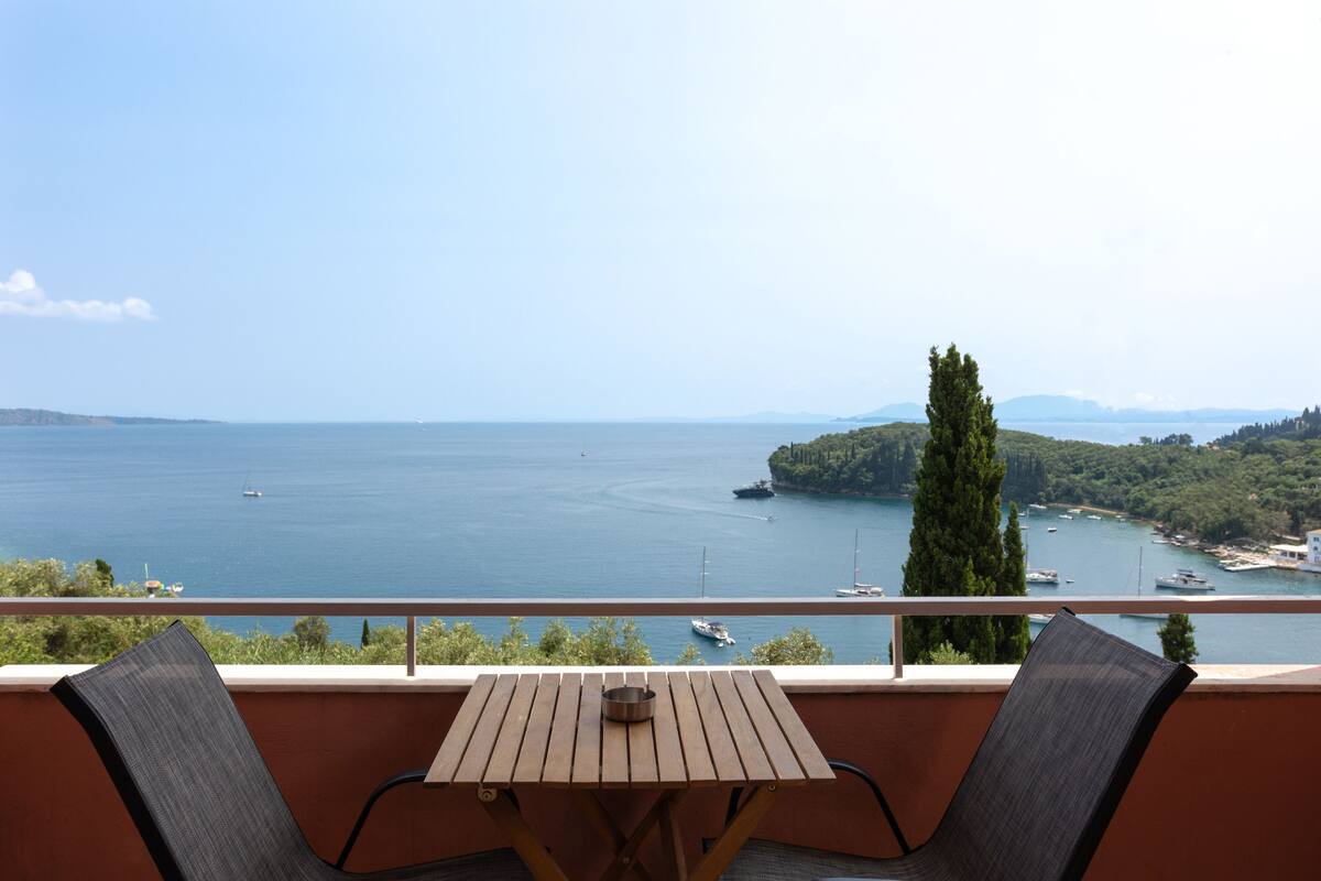 Ein Balkon mit Tisch und zwei Stühlen mit Blick auf ein Gewässer, wo Boote fahren und eine Insel im Hintergrund liegt.