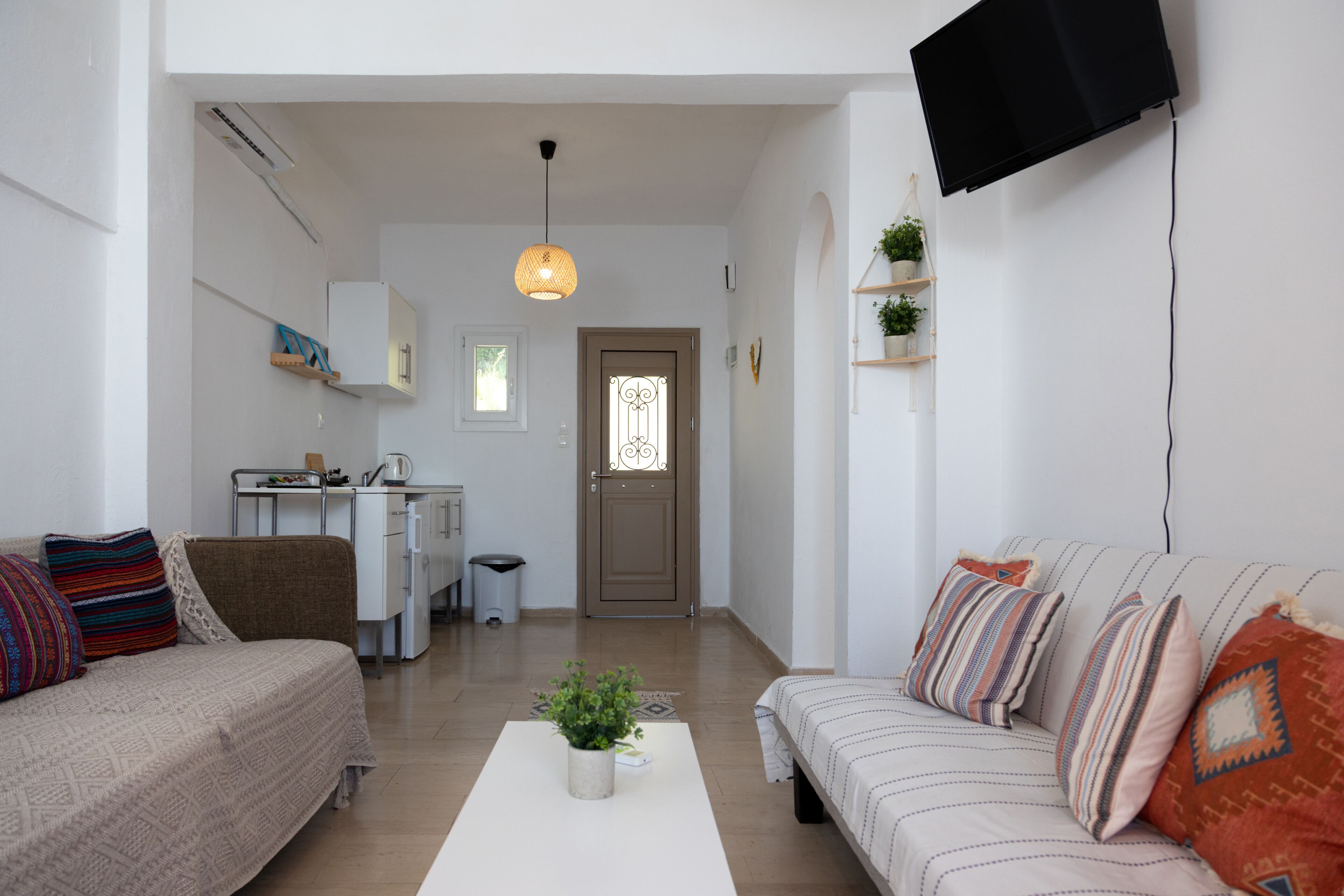 A modern living room with a sofa, a coffee table with a plant, a TV mounted on the wall, and a door leading to another room.