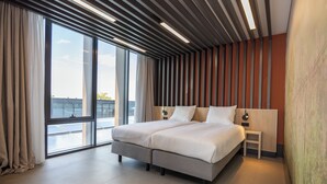 Una habitación de hotel moderna con una cama grande, una mesita de noche pequeña y vistas de la ciudad a través de amplias ventanas.