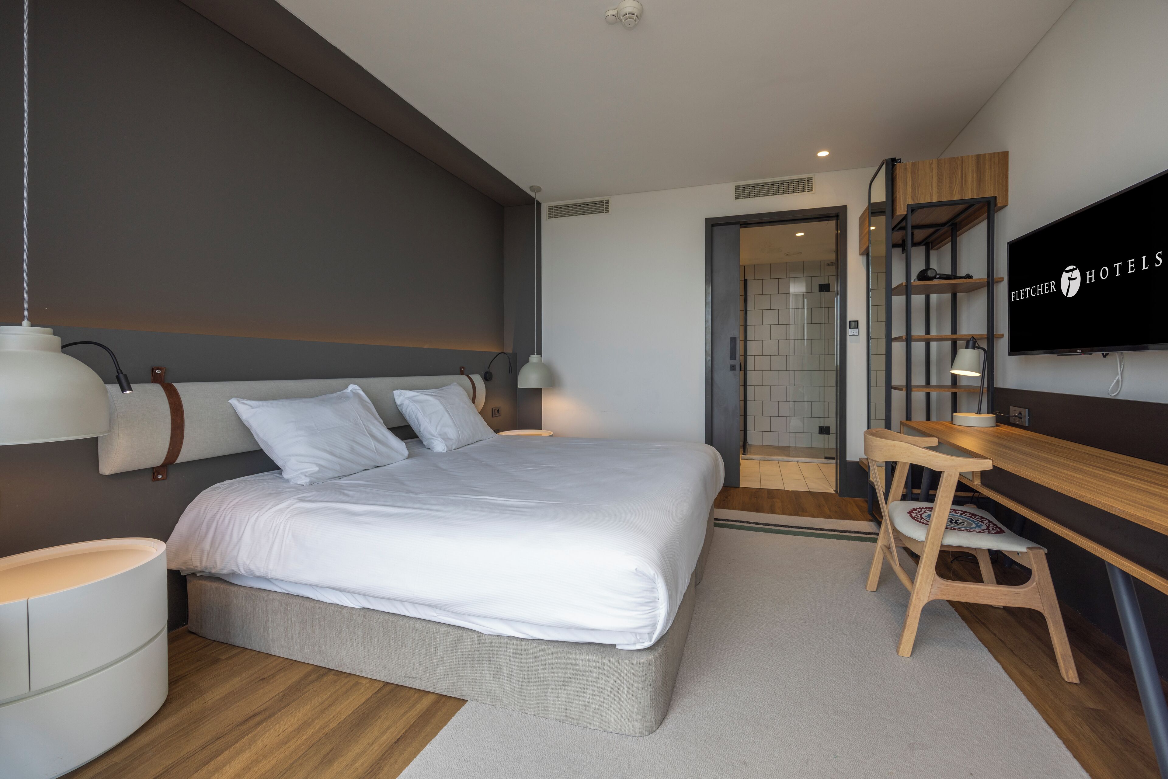 A modern hotel room with a large bed, bedside tables, a desk with a chair, a television, and a wardrobe.