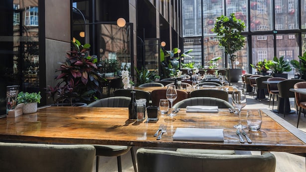 Un restaurante con una mesa de madera lista para comer, rodeada de sillas y amplios ventanales que ofrecen vistas de la ciudad.