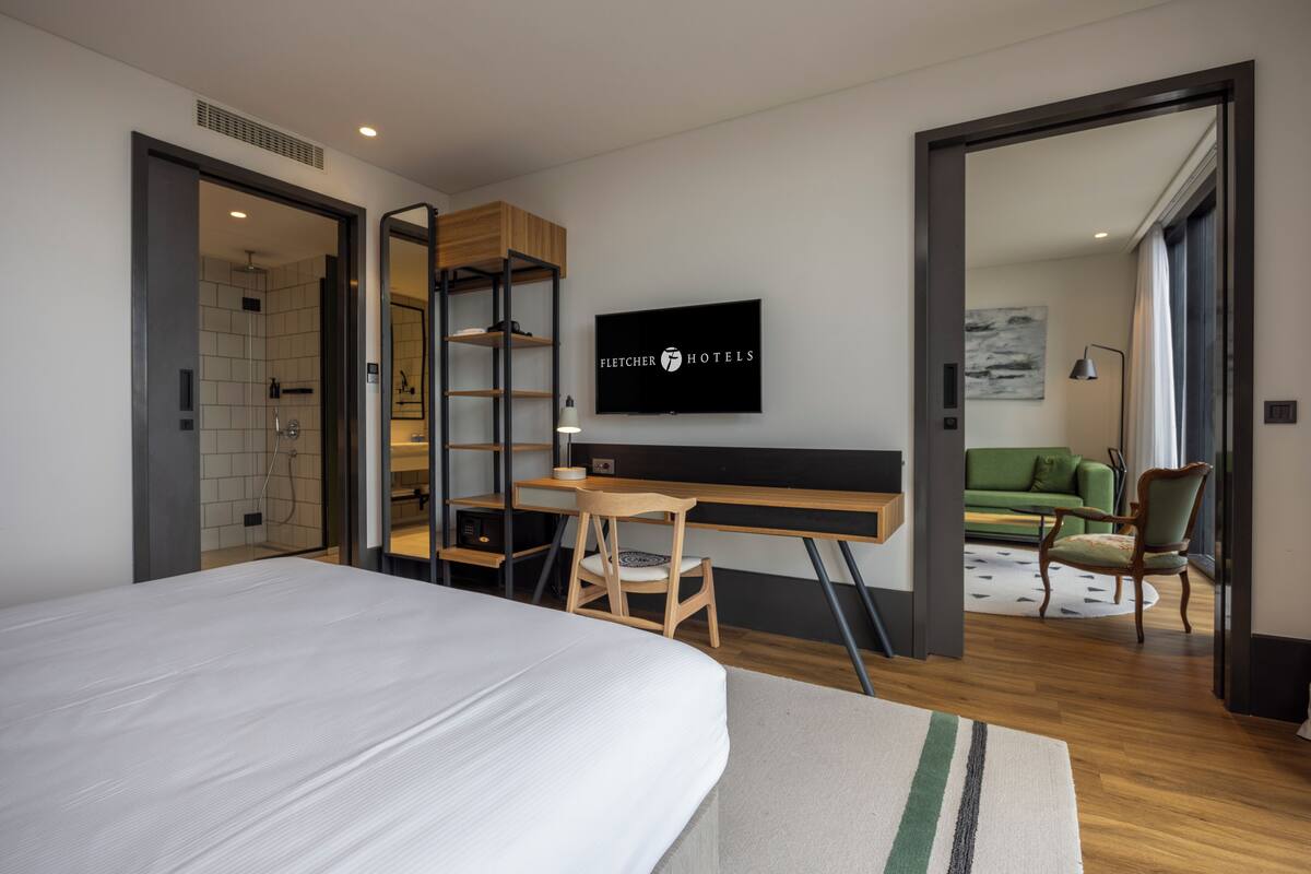A modern hotel room with a large bed, a desk with a chair, a TV mounted on the wall, and a view of a green sofa and a patterned rug in the adjacent room.