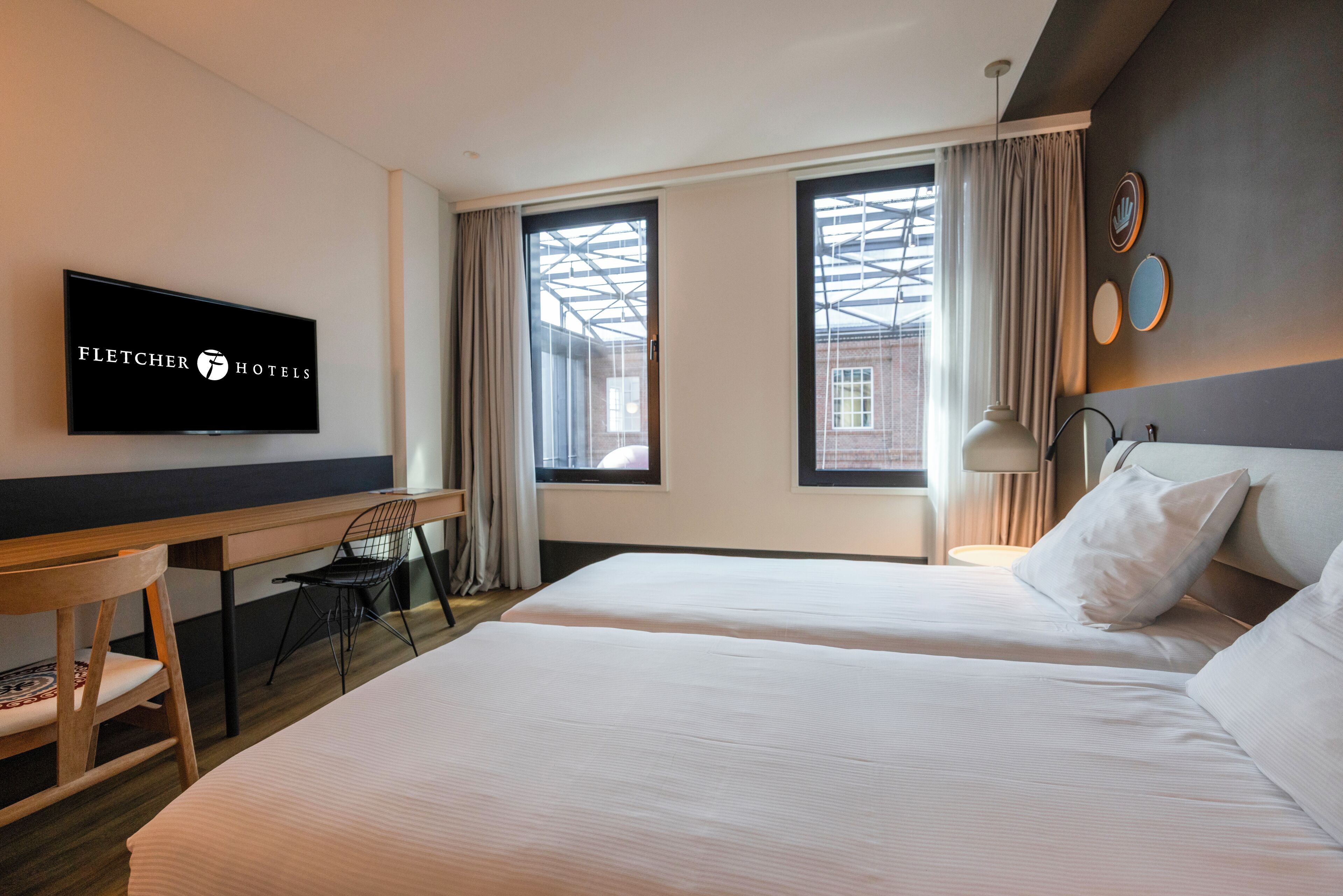 A hotel room with two beds, a desk, a chair, a TV, and large windows with curtains.
