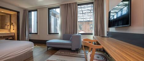 A modern hotel room with a bed, a desk, a chair, a TV, and large windows.