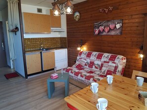 Ein gemütliches Blockhausszimmer mit einer Holzsofa, einem Esstisch mit weißen Tassen, einem kleinen blauen Tisch und einer Küchenzeile mit Holzschränken und einem Spülbecken.