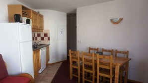 Cocina compacta con refrigerador, microondas y gabinetes de madera. Hay una mesa de comedor con sillas y un sofá rojo en la sala.