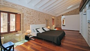 A bedroom with a stone wall, wooden flooring, a bed with a dark bedspread, bedside lamps, and a mirror on the wall. - Muralles - Central house with pool, air conditioning, and wifi in Alcúdia (Alcúdia)