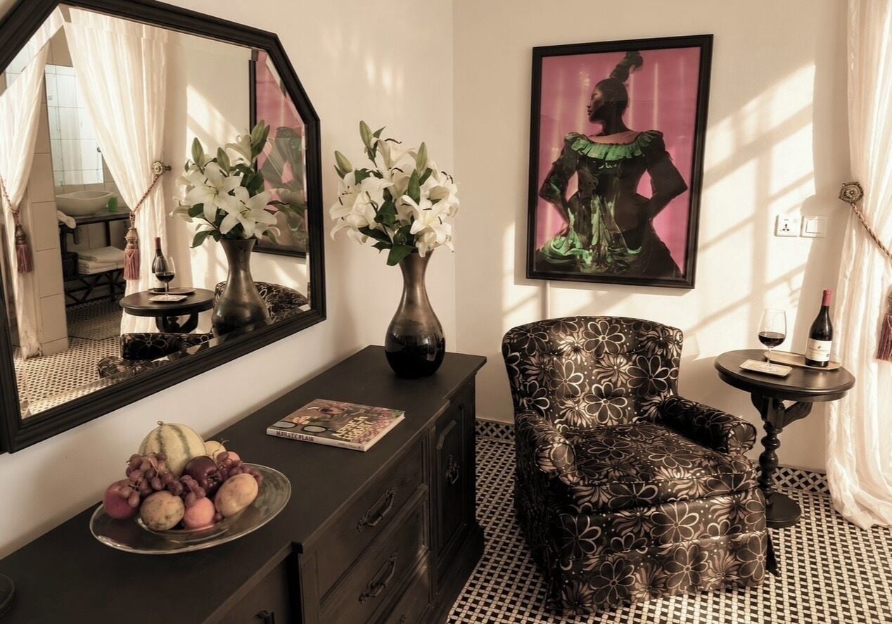 A room with a large mirror, a chair, a small table with a bottle, and a vase with flowers.