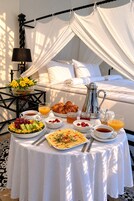 Un comedor exterior lujoso con una cama con dosel, una mesa con alimentos para el desayuno y una lámpara decorativa.