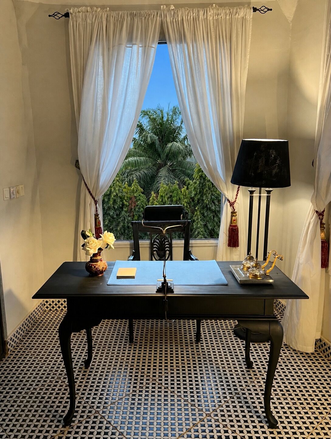 A room with a table, a lamp, a chair, a vase with flowers, and a view of palm trees through sheer curtains.