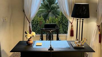 A room with a table, a lamp, a chair, a vase with flowers, and a view of palm trees through sheer curtains.
