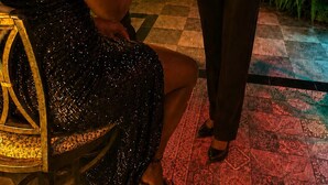 Una mujer con ropa negra está de pie junto a otra sentada con un vestido brillante, ambas en un entorno al aire libre con una palmera y una escalera de fondo.