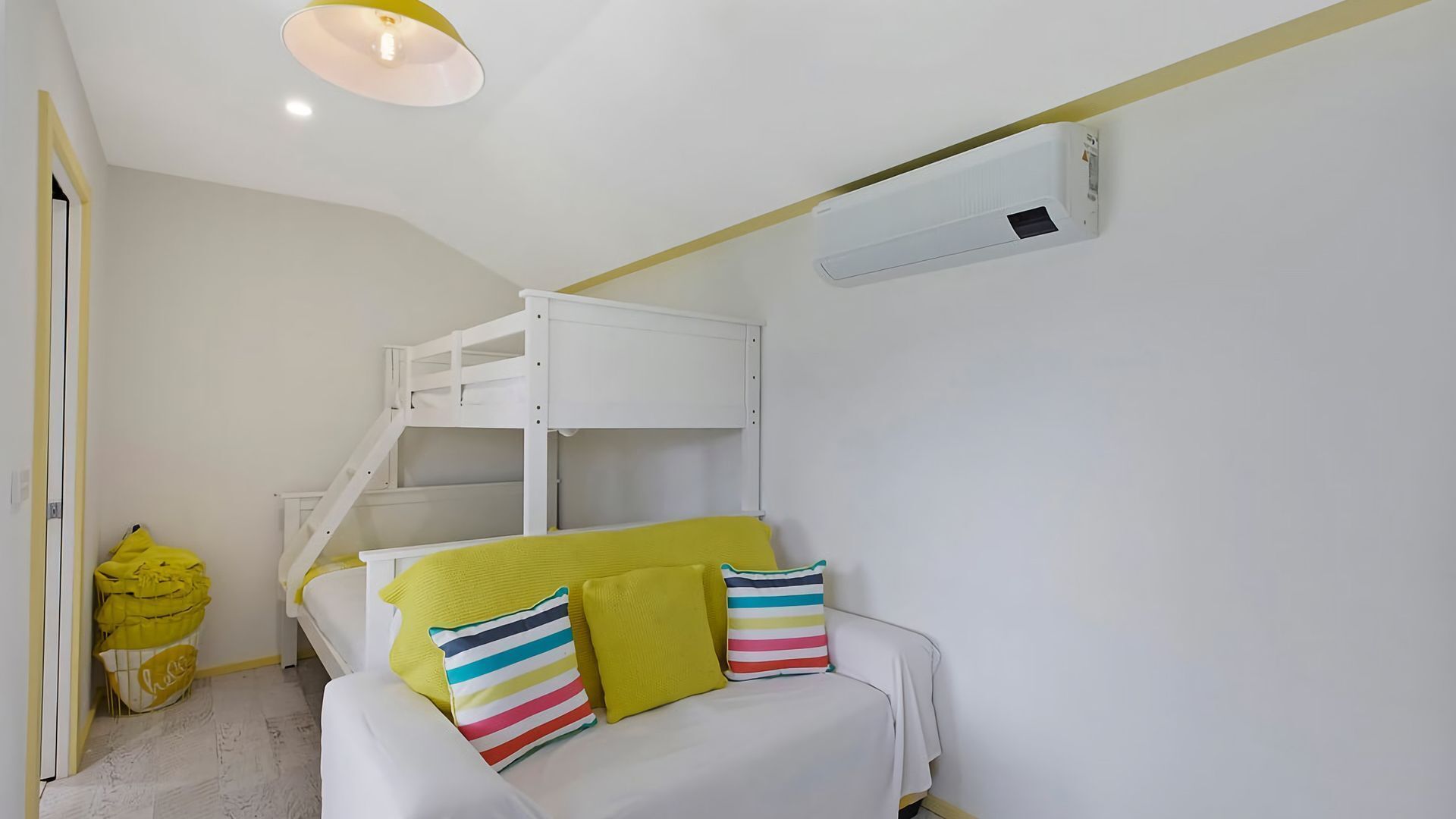 A room with bunk beds, a white sofa with colorful pillows, a yellow bag, and an air conditioning unit.