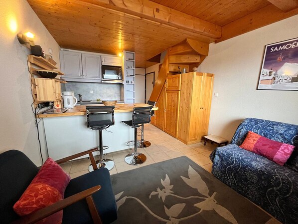 A compact living space with a kitchen, two bar stools, a sofa with patterned cushions, and a wooden ceiling.