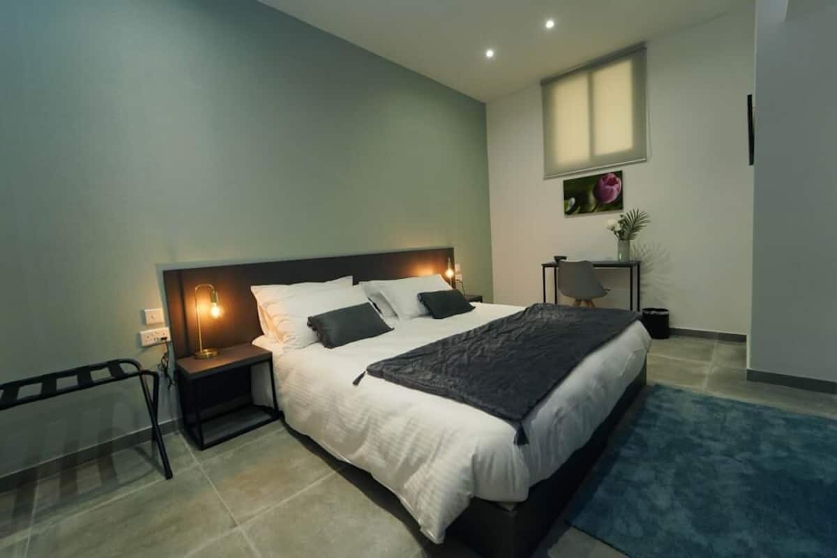 A modern bedroom with a large bed, bedside tables, a nightstand, a window, and a small table with a vase.