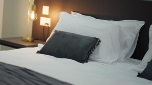 Cama bem arrumada com lençóis brancos e travesseiro cinza escuro, luminária de cabeceira e luminária fixada na parede.