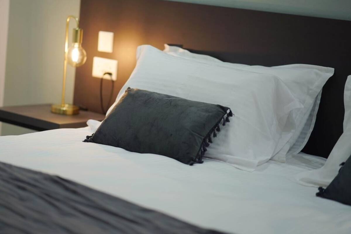 A neatly made bed with white linens and a dark gray pillow, a bedside lamp, and a wall-mounted light fixture.