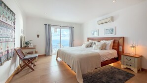A bedroom with a bed, bedside table, and a view of the sea through the window. - Villa Acacia- Spectacular Ocean view & heated pool (Arco Da Calheta)