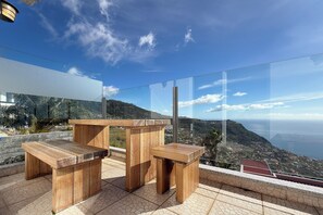 Eine Terrasse mit Holzmöbeln und Glasgeländer mit Blick auf eine Küstenlandschaft.