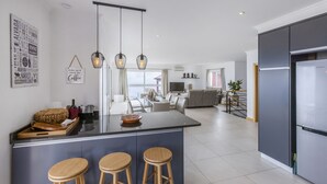 A modern kitchen with a central island, bar stools, and a dining area with a view. - Villa Acacia- Spectacular Ocean view & heated pool (Arco Da Calheta)