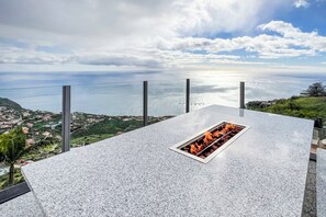 A stone patio with a built-in fire feature overlooking a coastal town.