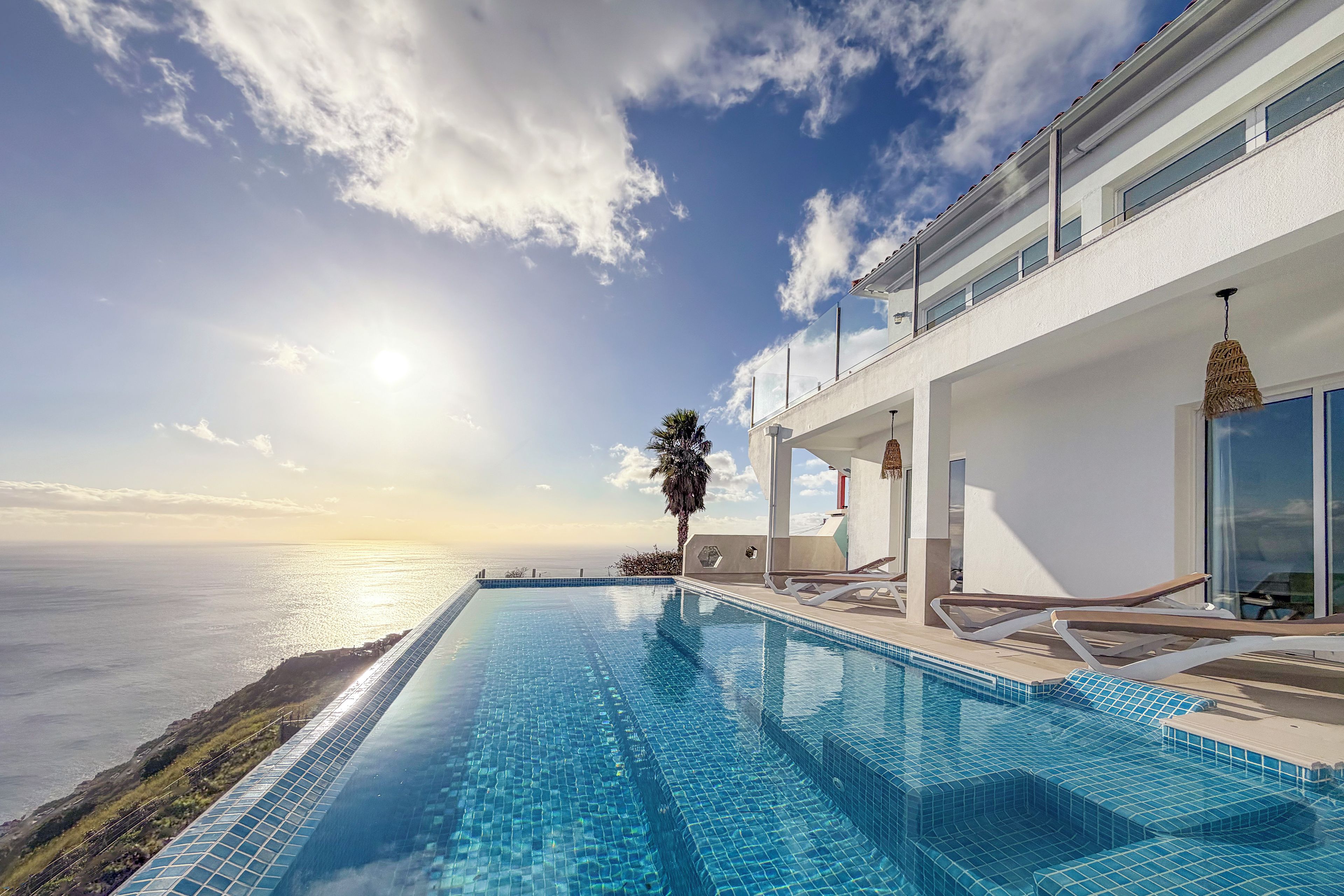 Une piscine à débordement moderne offrant une vue dégagée sur l’océan, un bâtiment blanc doté de balcons et un palmier.