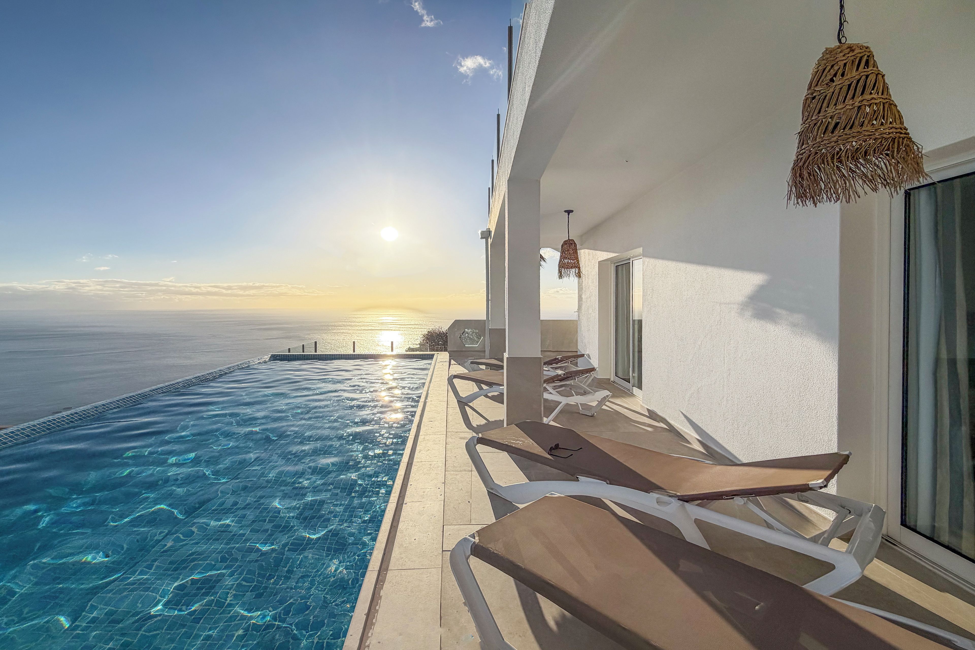 Un espace aménagé au bord de la piscine, avec vue sur l’océan au coucher du soleil, des chaises longues et un luminaire suspendu.