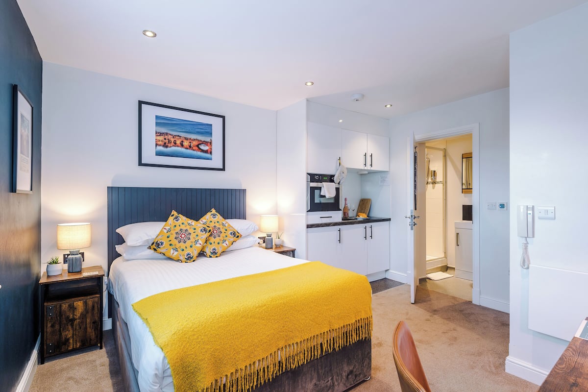 A hotel room with a bed, bedside tables, a desk, a chair, a kitchen area, and a bathroom.