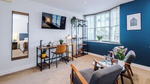 A modern room with a large mirror, a flat-screen TV, a desk with a chair, a small table with a vase of flowers, and a window with blinds. - Deva Leisure (Chester)