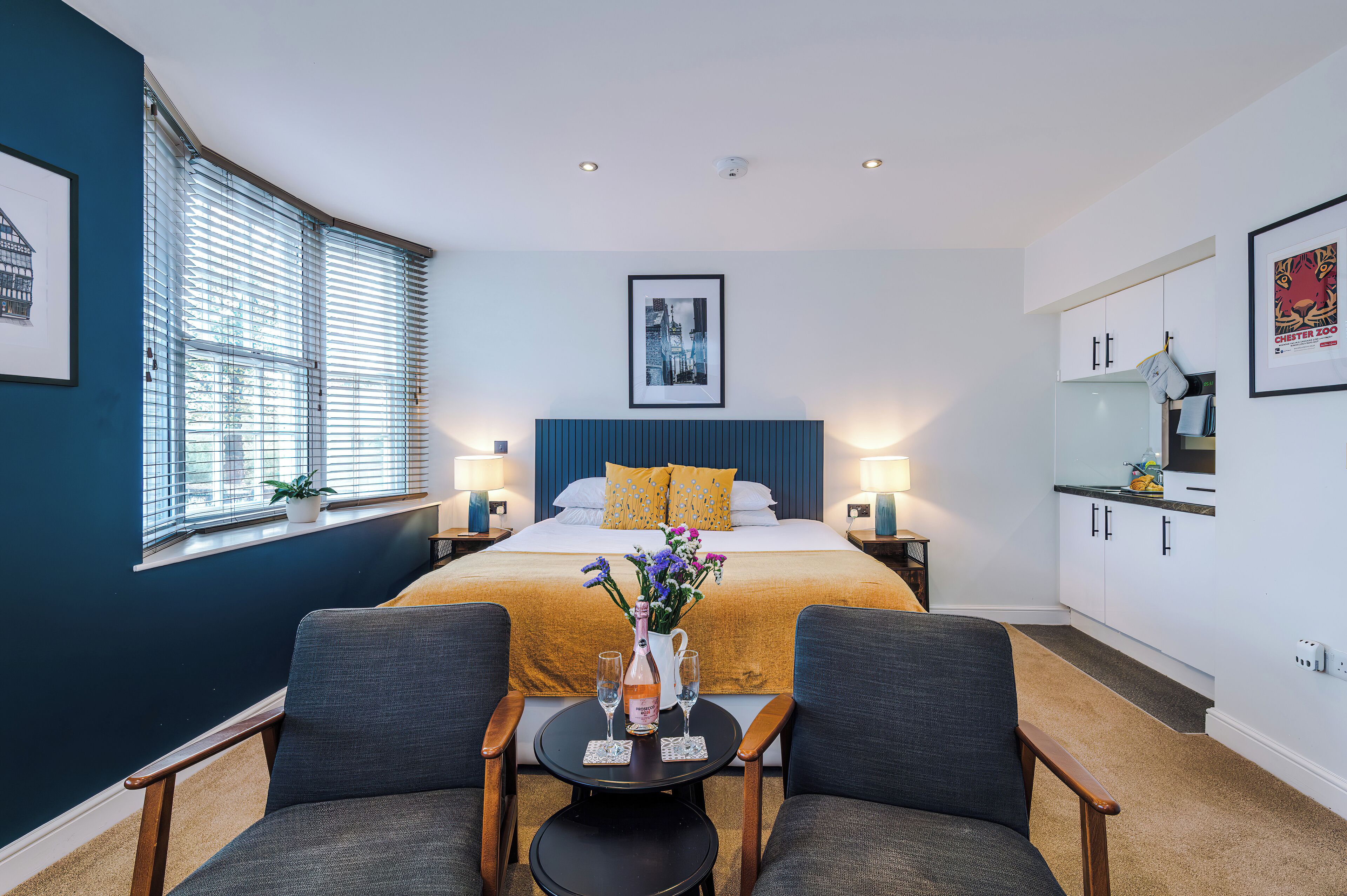 A hotel room with a large bed, a small table with a vase and glasses, two armchairs, a window with blinds, and a kitchenette.