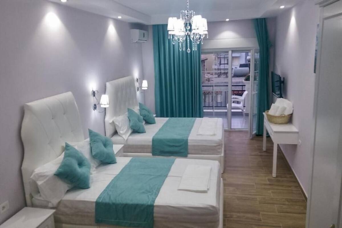 A hotel room with two beds, teal bedding, a chandelier, a small table with a basket, and a view of the balcony.