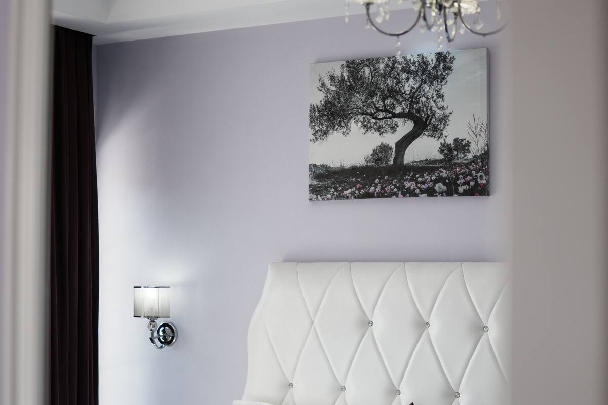 A bedroom with a tufted headboard, a bed with white linens and a purple throw, a wall-mounted lamp, and a painting of a tree.