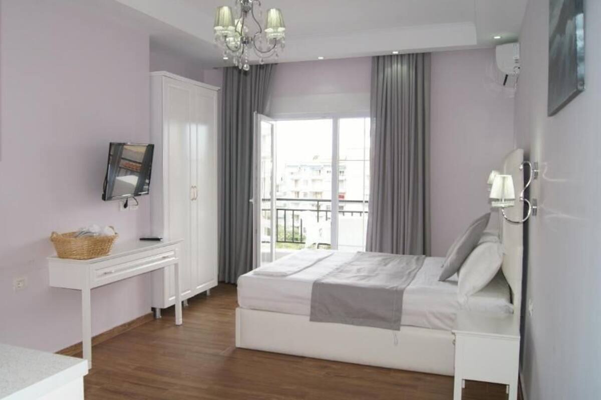 A bedroom with a bed, a desk, a chair, a chandelier, and a balcony with curtains.