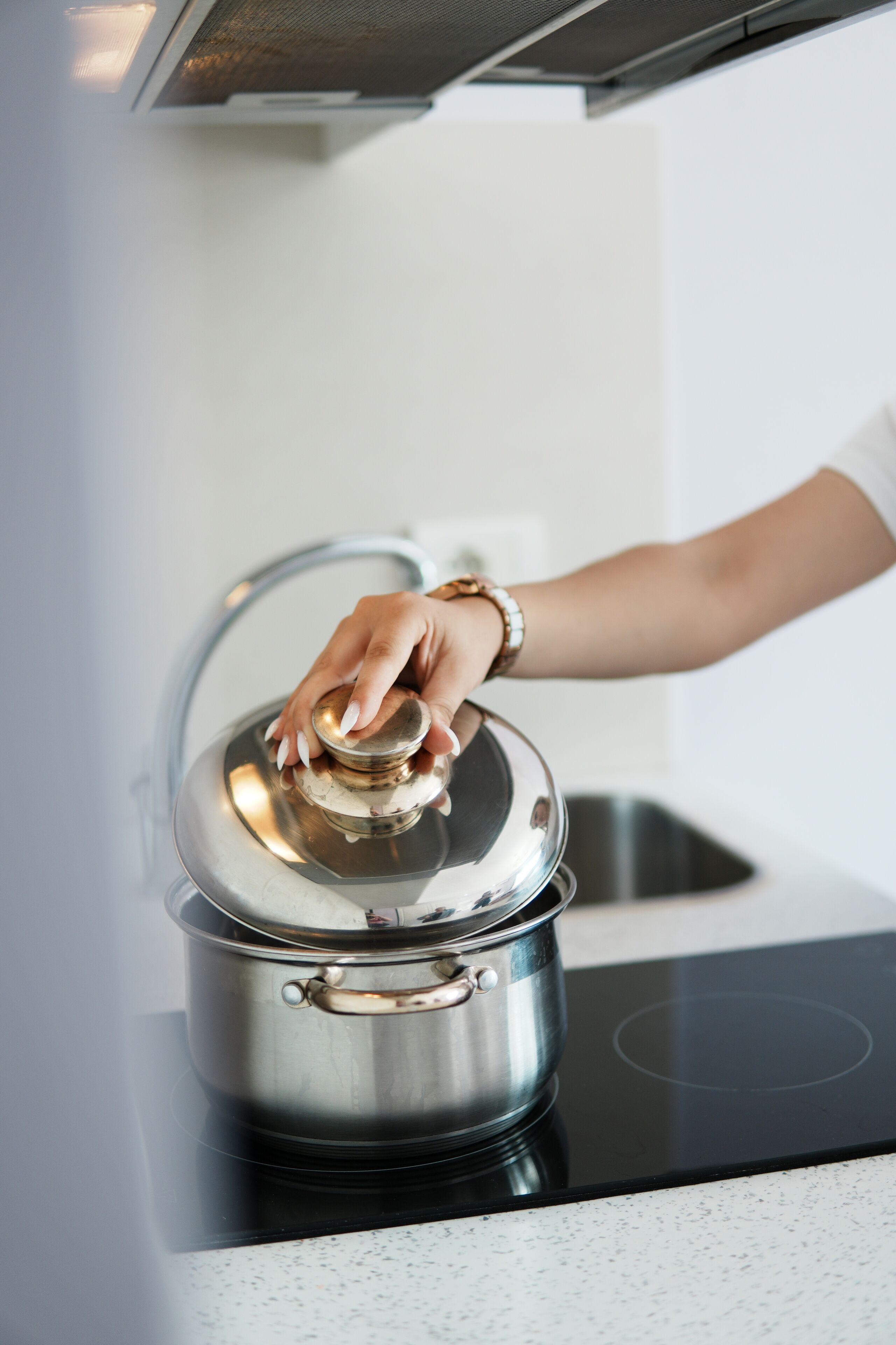 A hand lifting the lid of a stainless steel pot on an induction cooktop.