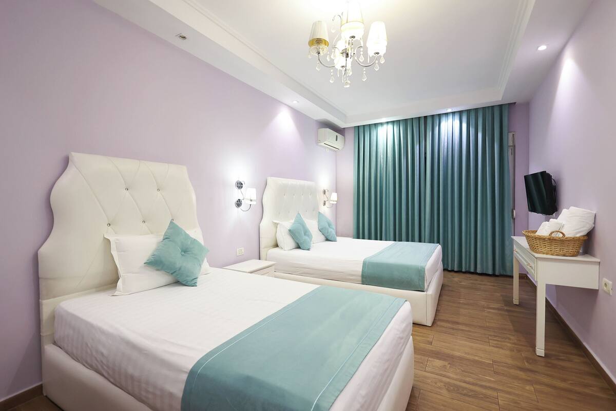 A hotel room with two beds, a chandelier, a small table with a basket, and a TV on a stand.
