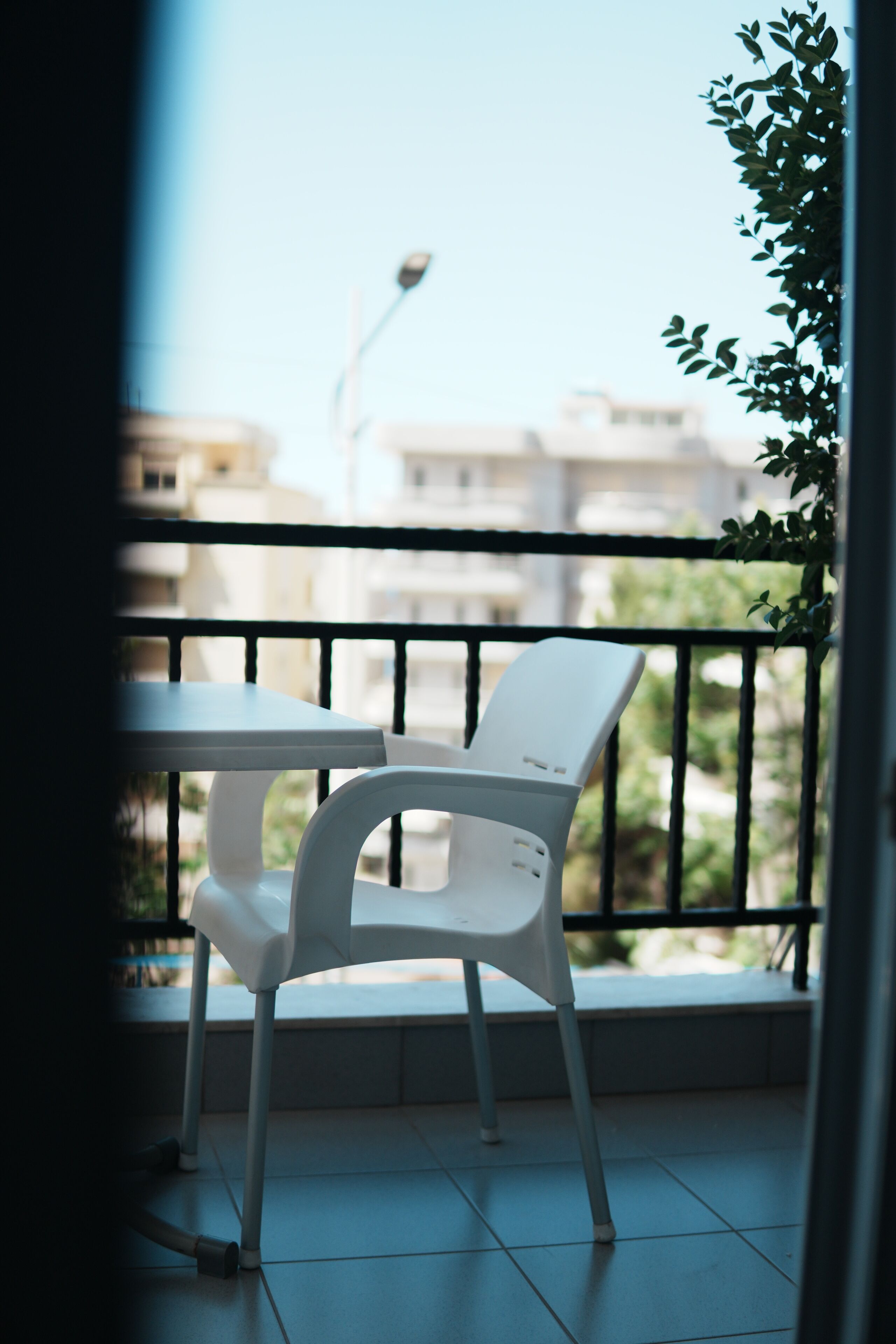 Un balcón con una silla y una mesa blancos, barandilla y vistas a edificios y follaje.