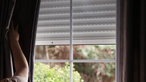 A window with blinds partially drawn, a person reaching to adjust them, and a view of greenery outside. - Apartments Flower Residence (Sarandë)