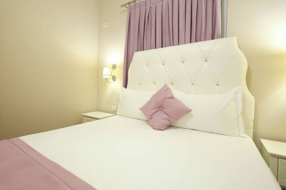 A neatly made bed with a tufted headboard, a single pink pillow, and a white bedspread.