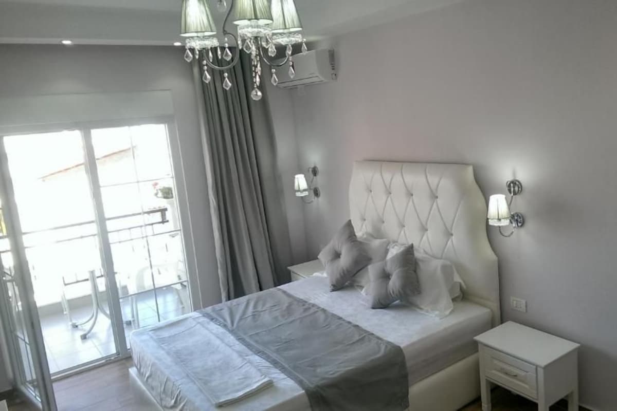 A bedroom with a large bed, a bedside table, a nightstand, a chandelier, and a painting on the wall.