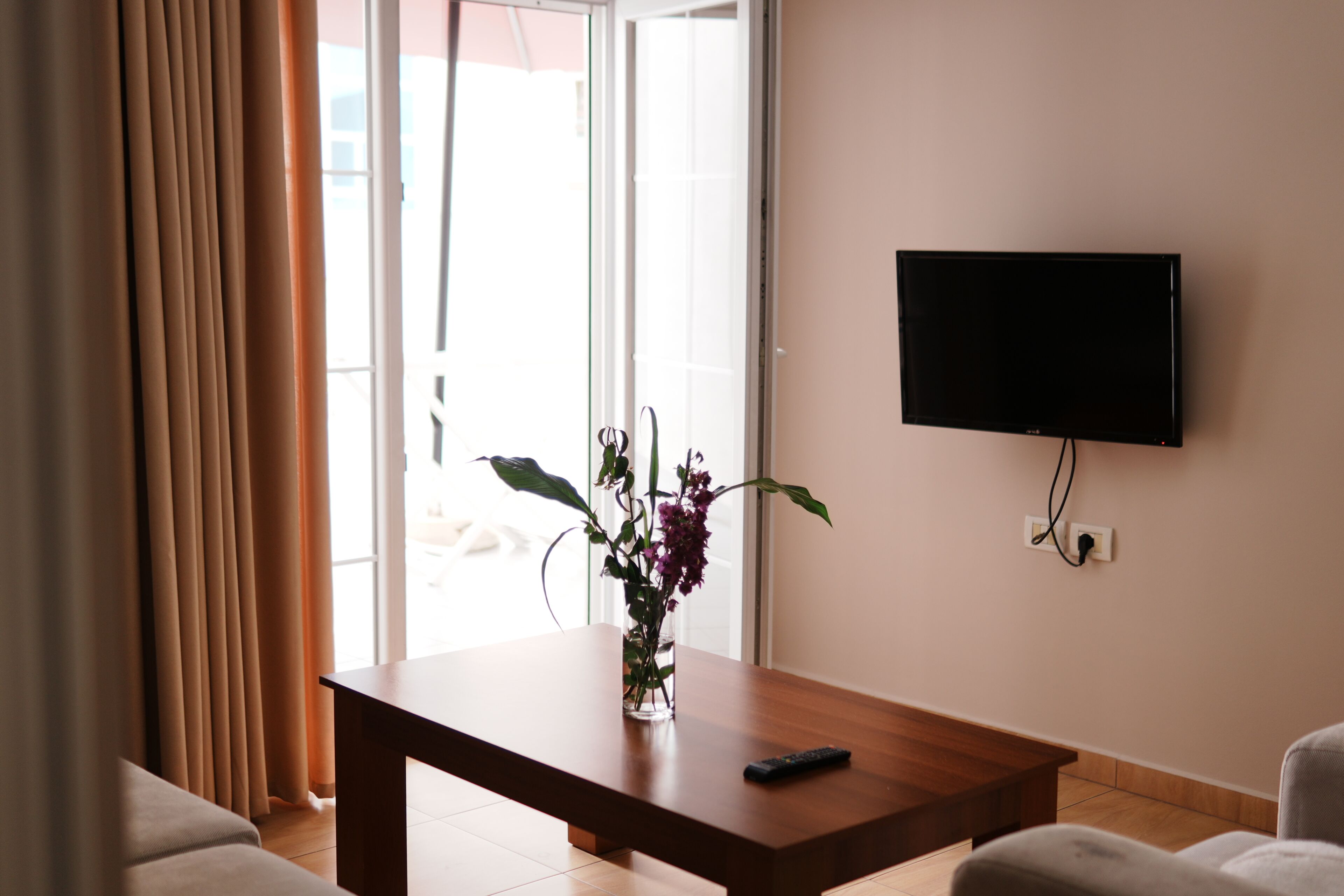 Une pièce avec une table en bois, un vase de fleurs, une télécommande, un téléviseur fixé au mur et une porte-fenêtre coulissante.