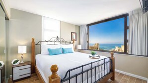 Een slaapkamer met een bed, nachtkastjes, een raam met uitzicht op zee, en een balkon.