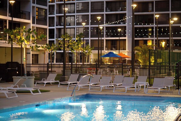 Un área de piscina con sillones de descanso, una piscina cercada y un edificio con varios pisos y balcones.