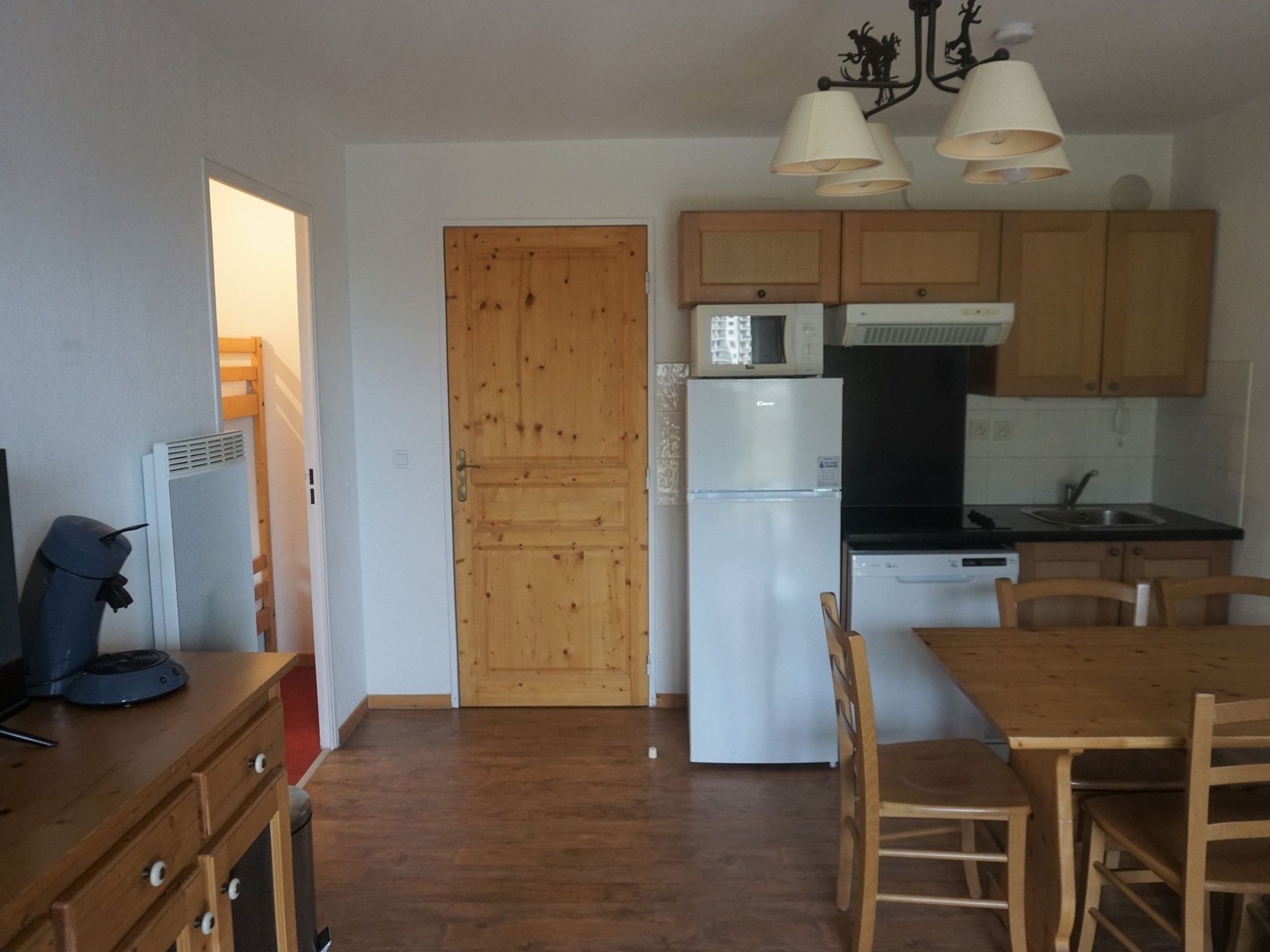 Une cuisine compacte avec des meubles en bois, un réfrigérateur blanc et un four à micro-ondes. On y trouve une table à manger avec des chaises et un luminaire au plafond.
