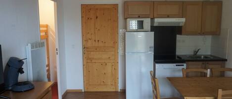 A compact kitchen with wooden cabinets, a white refrigerator, and a microwave. There is a dining table with chairs and a ceiling light fixture.
