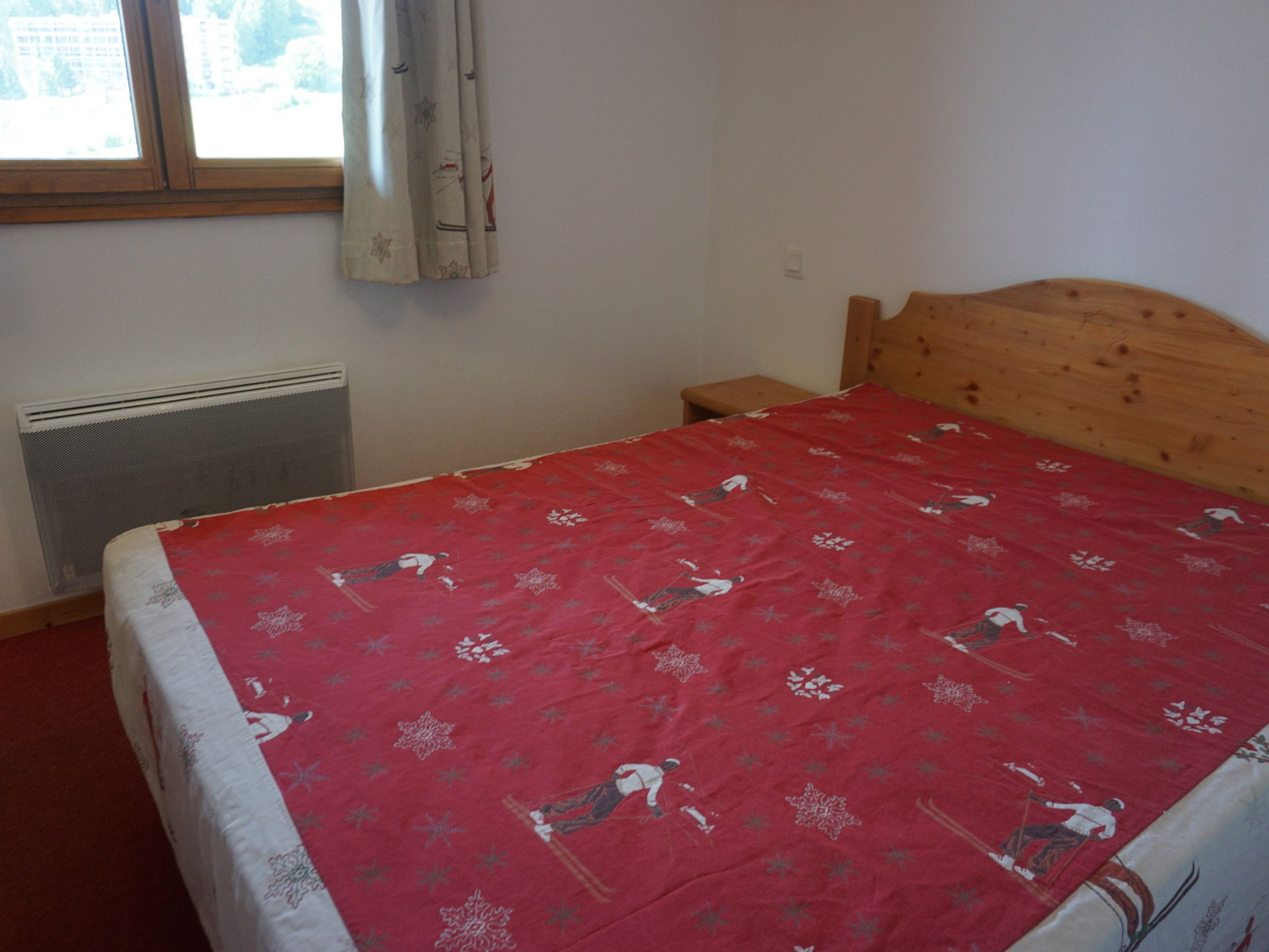 A bedroom with a wooden headboard, a bed with a red and white patterned bedspread, a small bedside table, and a window with curtains.
