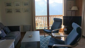 Un salon comprenant un canapé, deux fauteuils, une table basse et un téléviseur. Des tableaux encadrés ornent le mur, et la fenêtre offre une vue sur les montagnes.
