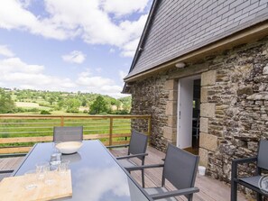 A stone building with a wooden deck, a table with chairs, and a scenic view of green hills.