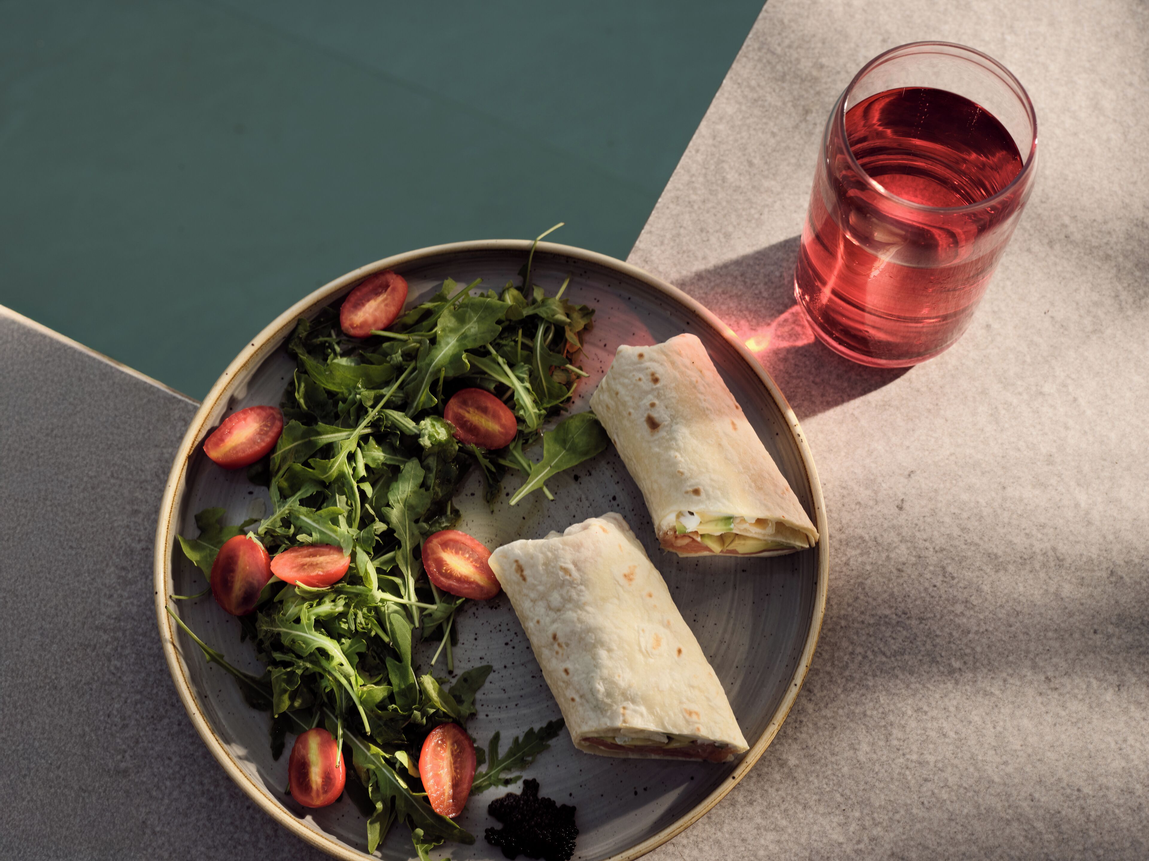 A plate with a salad and two wraps, and a glass of pink drink on a table by a pool.