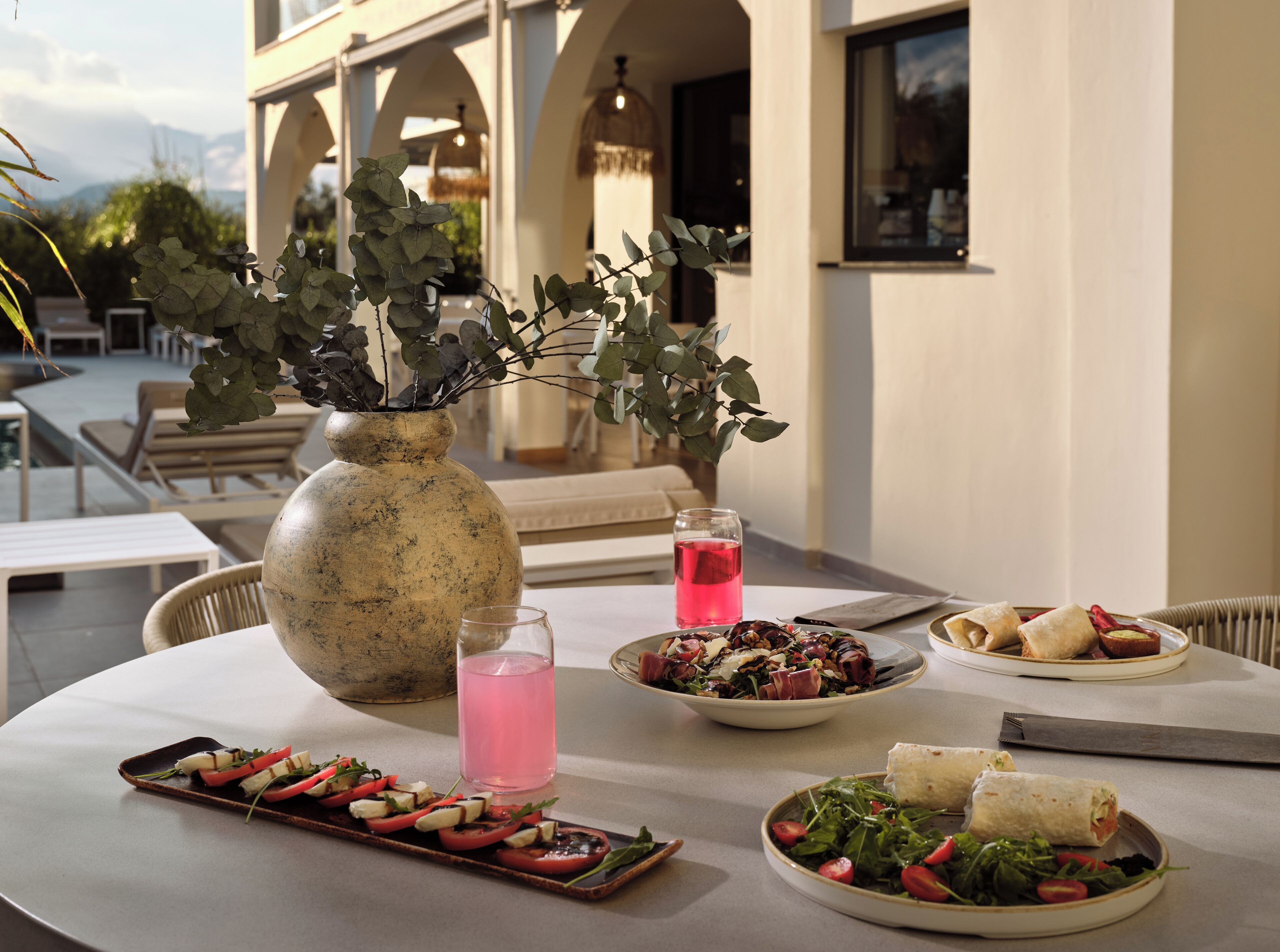 A table set with food, a vase with greenery, and a glass of pink drink.