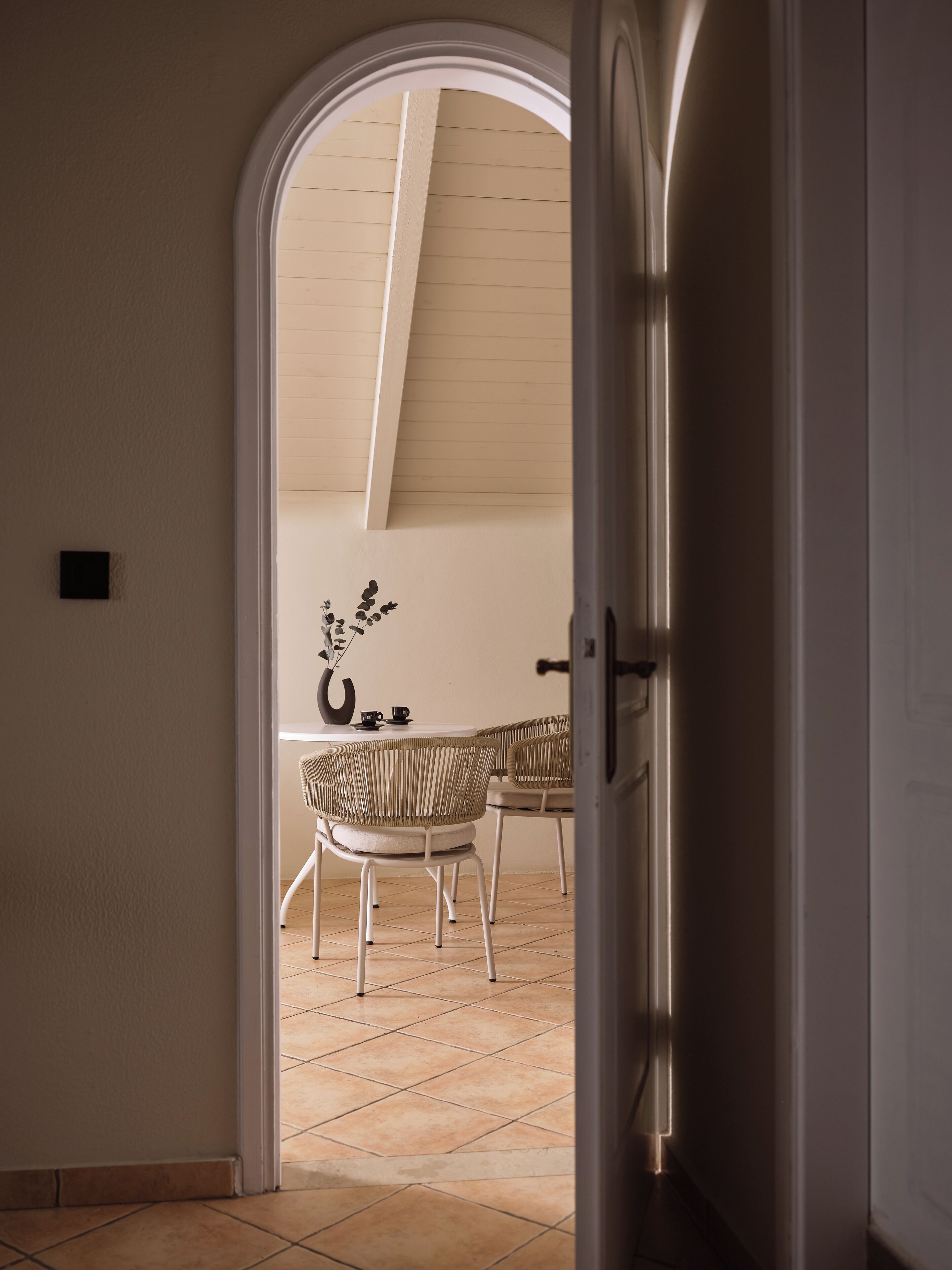 Een zicht in een kamer met een hoge plafond, een eethoek met rieten stoelen en een tafel, en een deur die naar een andere ruimte leidt.